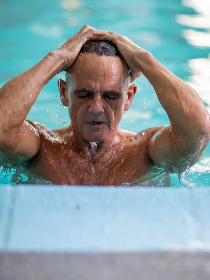 Client surfacing at a pool wall, pushing soaking wet hair straight back off his forehead with both hands