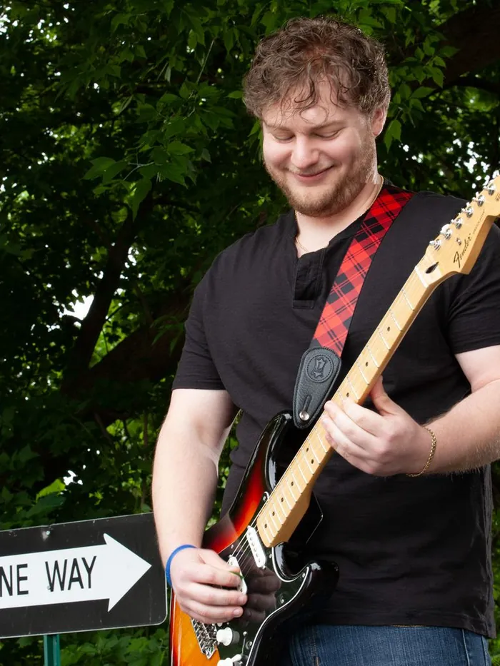 Client playing an electric guitar outdoors on a street corner in full sunlight