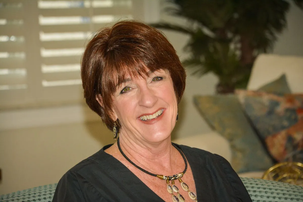 Client with a short auburn layered hair system smiling while seated in a bright sunroom