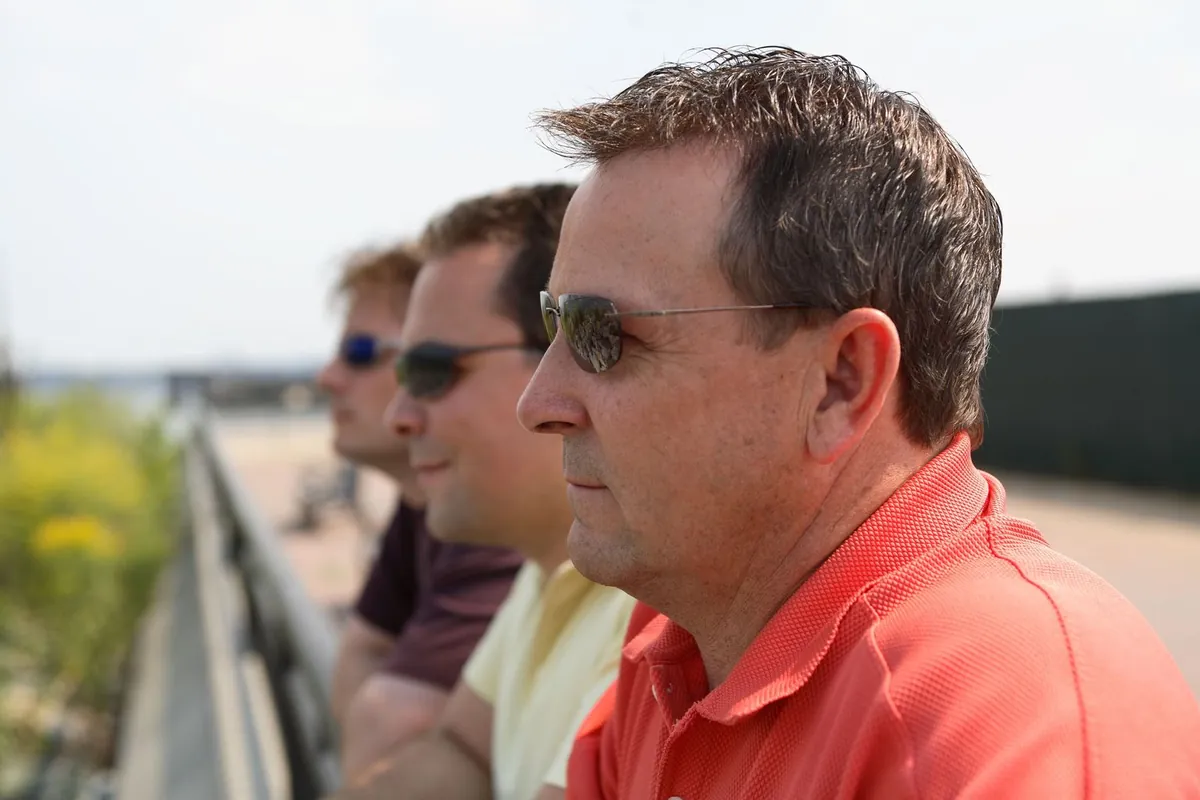 Man in his fifties in a red polo and sunglasses, seen in profile outdoors with two friends on a waterfront walkway